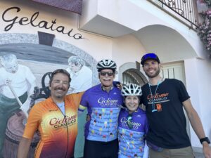 Group of four cyclists in Italy wearing cycling gear, smiling, with a mural and gelato shop in the background, promoting Italy bike tours and top bike tour companies.
